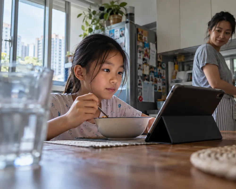 A girl eats breakfast in front of a tablet while her mother cooks in the background